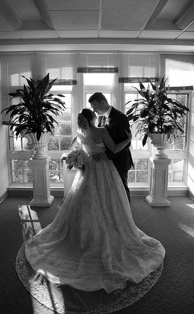 A bride and groom lovingly embrace, framed by a grand window. Their joyous union radiates through their tender connection.