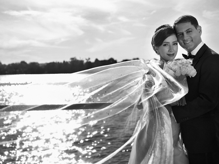 A black and white photo captures a bride and groom posing by the water, radiating love and joy on their special day.