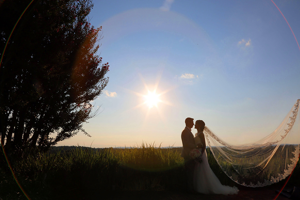 Couple stands together, smiling, in front of a radiant sun on their wedding day.