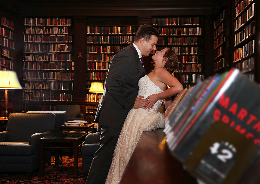Couple in wedding attire stands among bookshelves, radiating love and joy on their special day