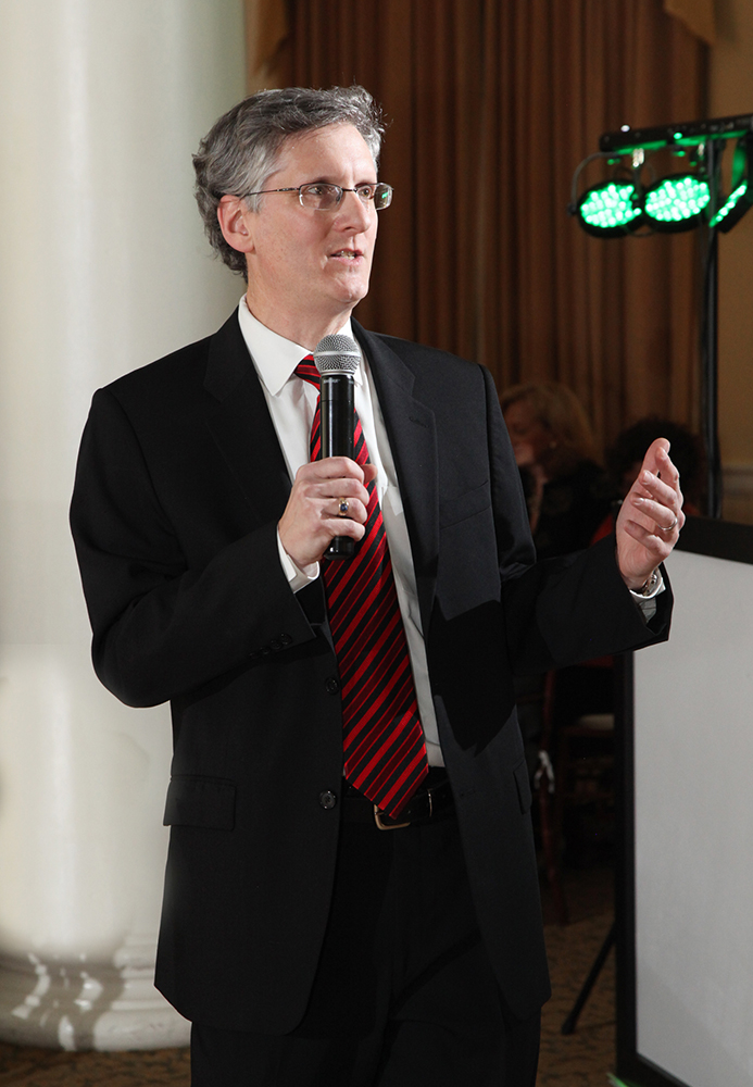 A confident professional man in a suit and tie, holding a microphone, ready to address an audience.