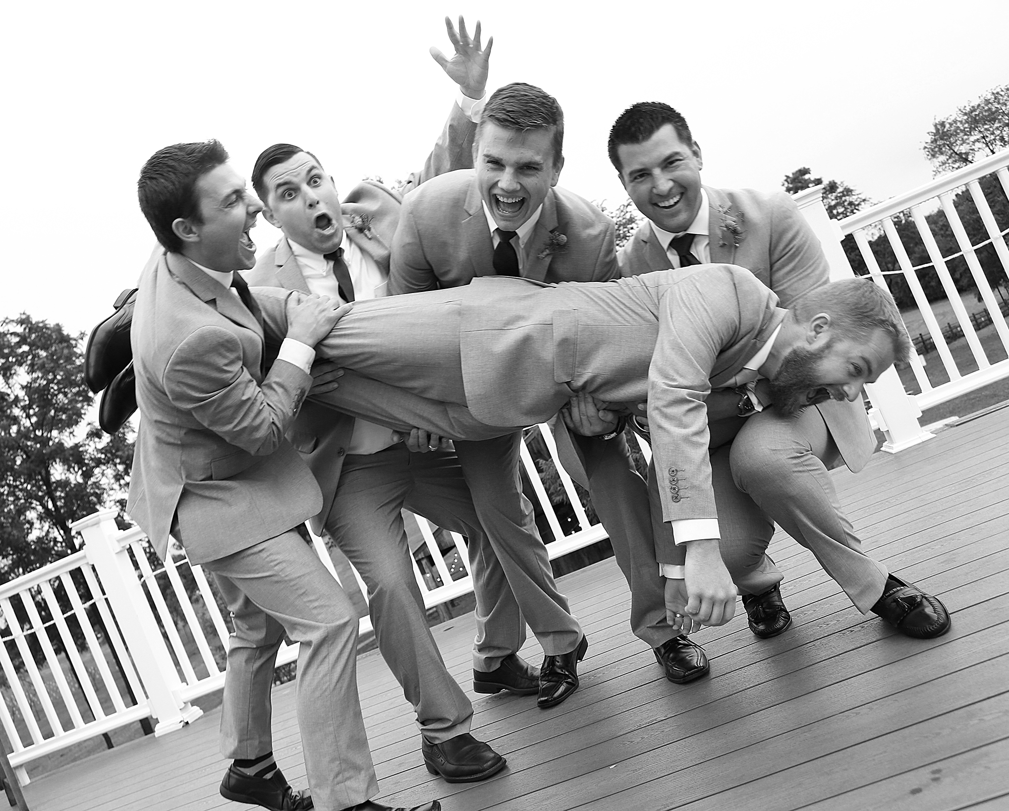 Groomsmen joyfully lift the radiant bride on a deck, their faces alight with happiness and celebration in this delightful and spirited moment.