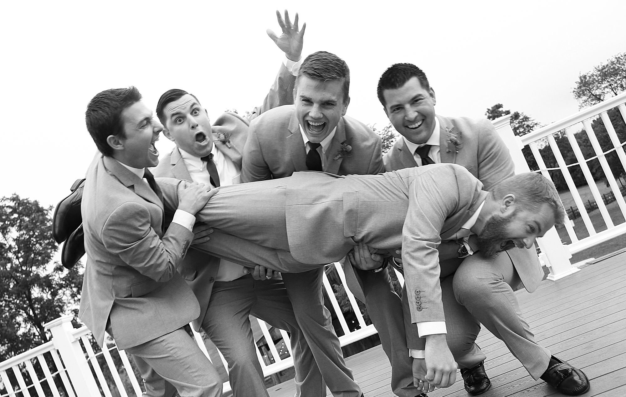 Groomsmen joyfully lift the radiant bride on a deck, their faces alight with happiness and celebration in this delightful and spirited moment.