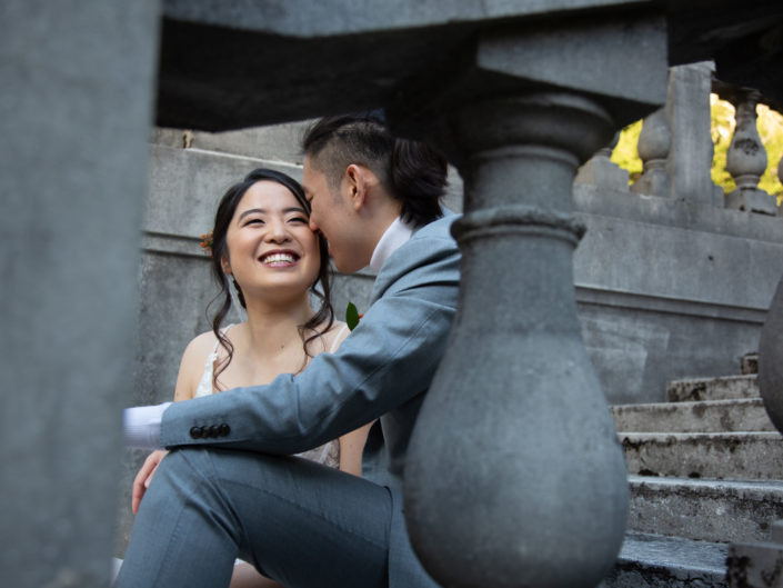 A couple, dressed in wedding attire, sits on steps outside a building, radiating joy and love on their special day.
