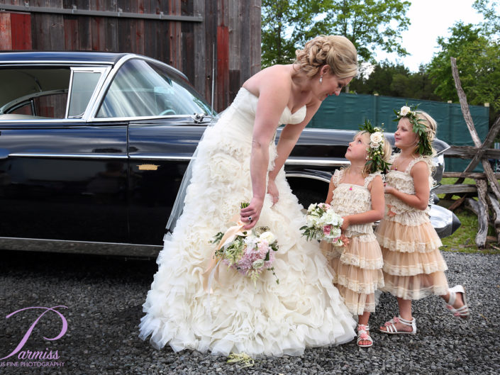 Bride and flower girls pose gracefully by vintage car.