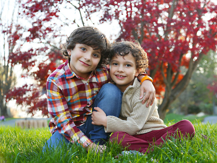 Two boys sitting on the grass in front of a red tree.